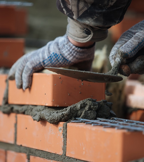 Mains gantées d'un maçon posant une brique sur du mortier frais, avec une truelle étalant le ciment sur un mur en construction.