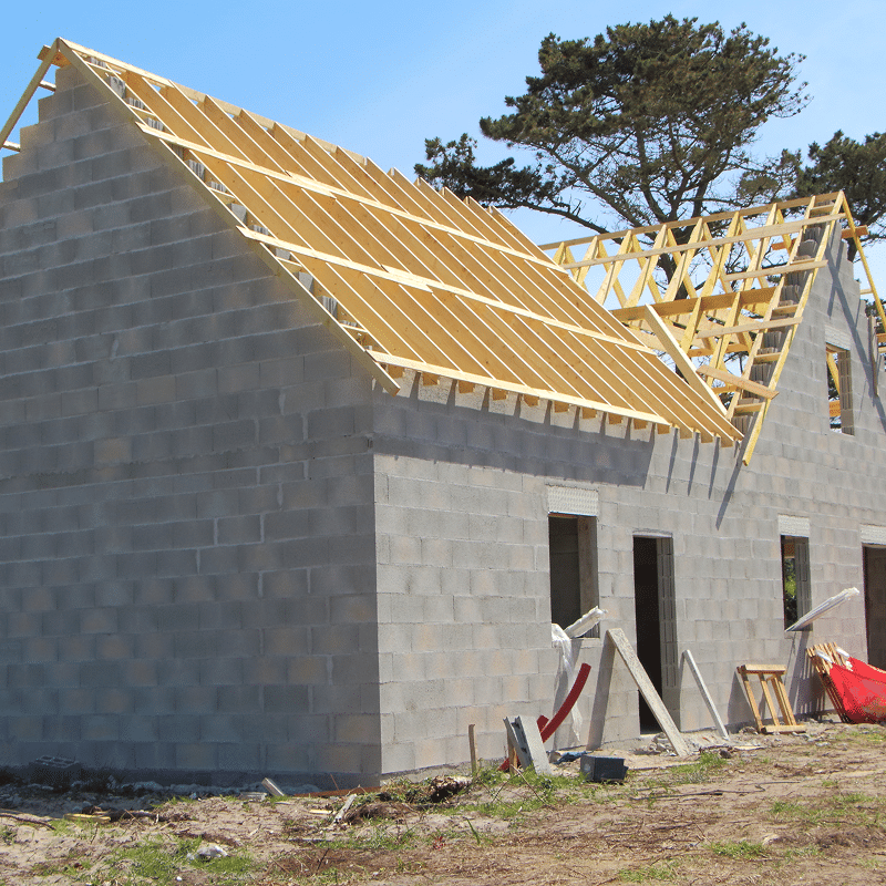 Maison neuve en parpaings gris, charpente de toit en bois clair en cours de pose sous ciel bleu. Chantier de construction.
