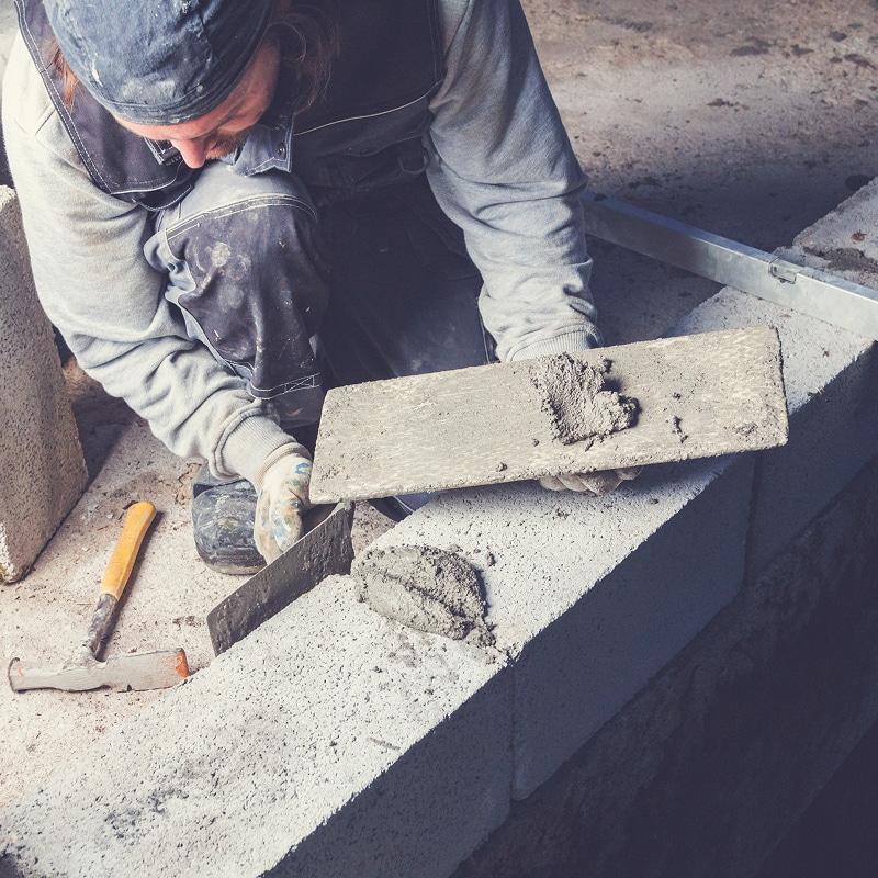 Un maçon applique du mortier sur des blocs de béton à l'aide d'une truelle et d'une taloche pour construire un mur. Outils visibles.