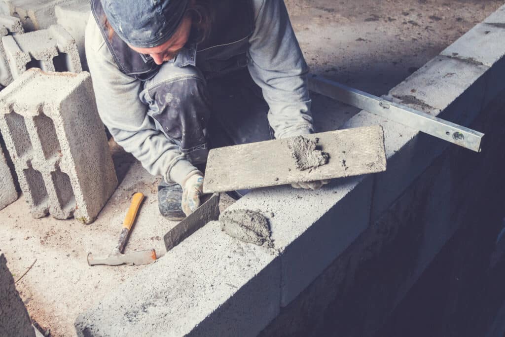 Un bâtisseur pose des parpaings en béton avec du mortier et une truelle, un niveau de maçonnerie et d'autres blocs sont sur le chantier.