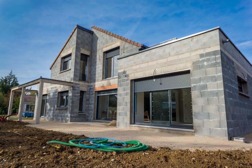 Maison moderne en parpaings en construction, fenêtres et volets installés. Tuyaux au premier plan sur le chantier sous ciel bleu.