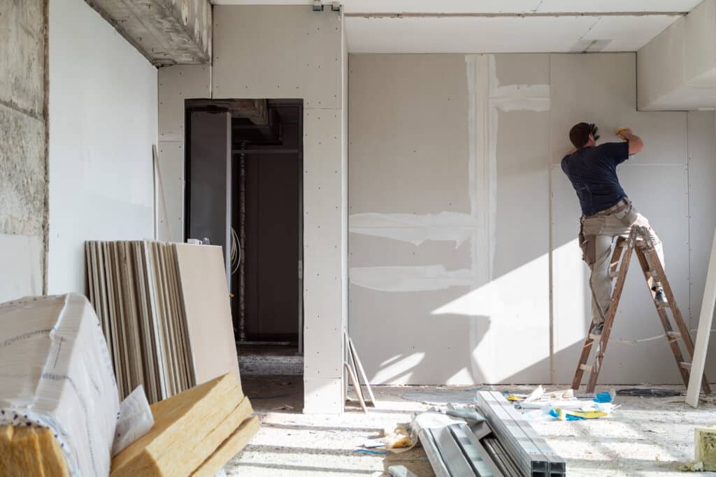 Ouvrier sur une échelle fixant une cloison sèche dans un intérieur en rénovation. Matériaux de construction, comme le placo et l'isolation, sont au sol.