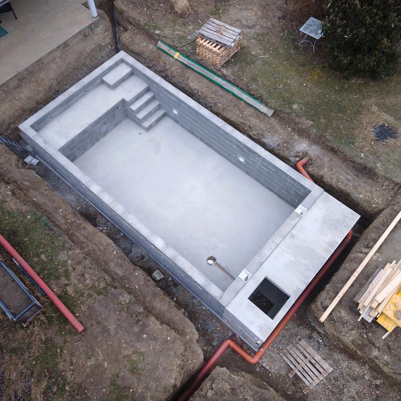 Piscine en béton brut en construction dans une excavation. La coque grise a des marches intégrées, entourée de tuyaux de drainage.