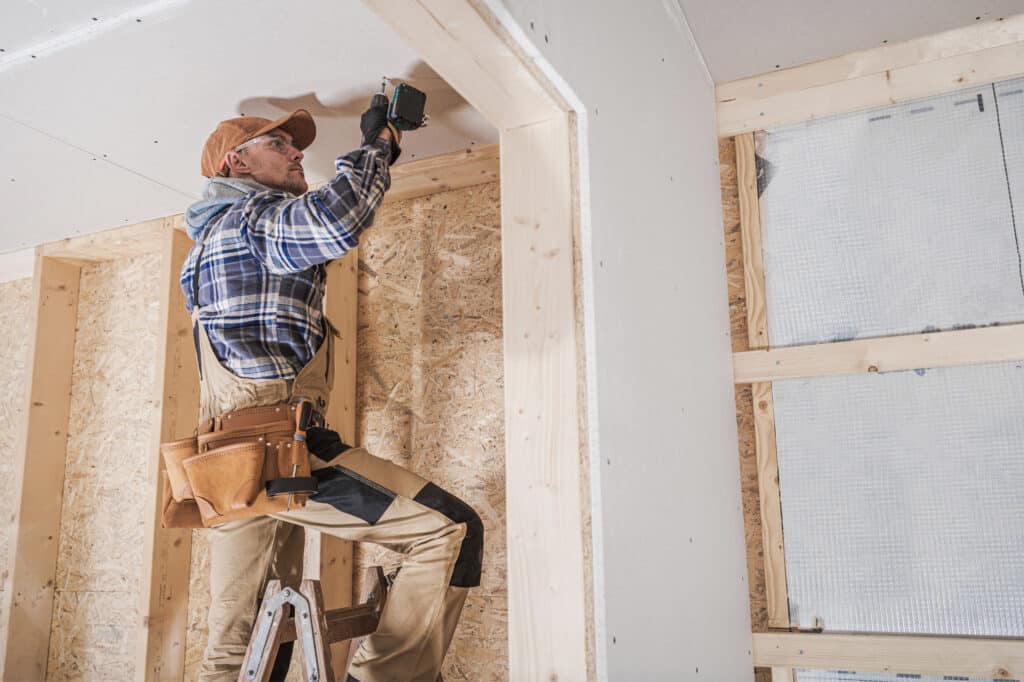 Homme sur échelle fixant du placo au plafond avec une visseuse. Casquette, lunettes de sécurité et ceinture porte-outils. Chantier bois OSB.